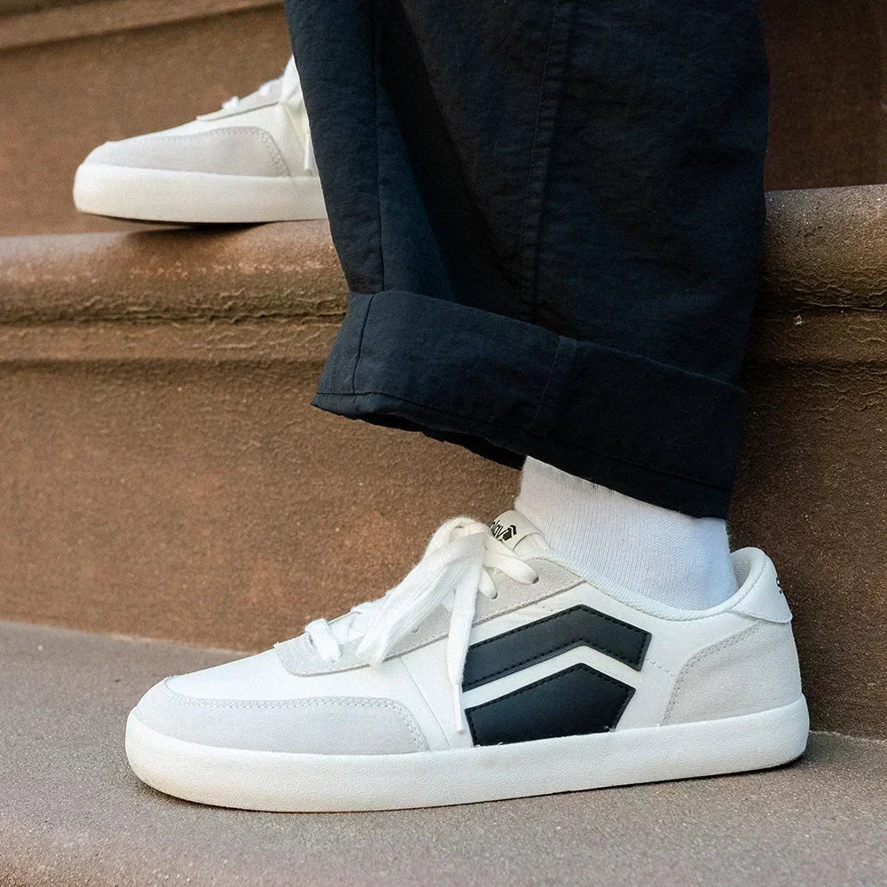 Close-up of white and black Splay barefoot sneakers worn with cuffed black pants while sitting on outdoor steps.